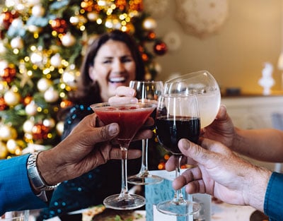 Four hands raise various cocktails in a toast, with a festive Christmas tree adorned with lights and ornaments in the background.