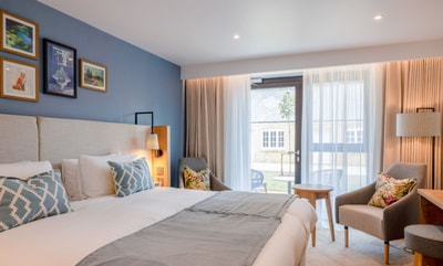 New Deluxe room at Heythrop Park Hotel with a king-sized bed, plush pillows, decorative art, and chairs beside a window overlooking a garden patio.