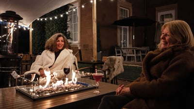 Two women sat in fur jackets by a firepit and enjoying cocktails together