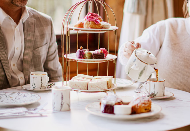 Tea being poured from a teapot into a teacup next to a tiered stand of sandwiches, cakes, and macarons, with plates of scones and jam arranged on the table.