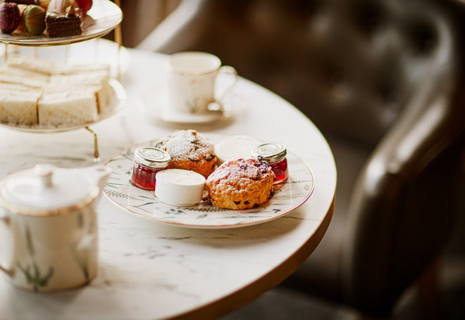 Close-up of a plate with scones dusted with icing sugar, served with small jars of jam and clotted cream on a marble table, with teacups and a tiered afternoon tea stand softly blurred in the background.