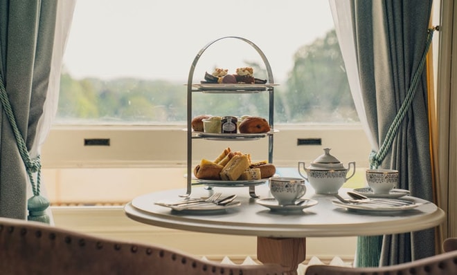 A charming afternoon tea set with an elegant three-tier stand filled with pastries, beside fine china cups and a teapot, against a scenic window view.