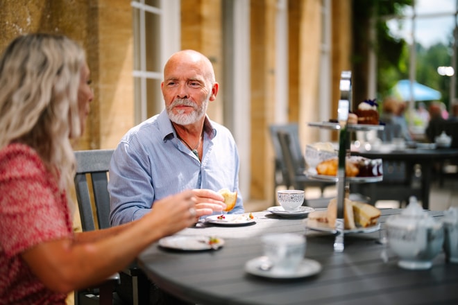 A couple enjoys afternoon tea outdoors, with plates of pastries and a tea set on a wooden table surrounded by greenery.