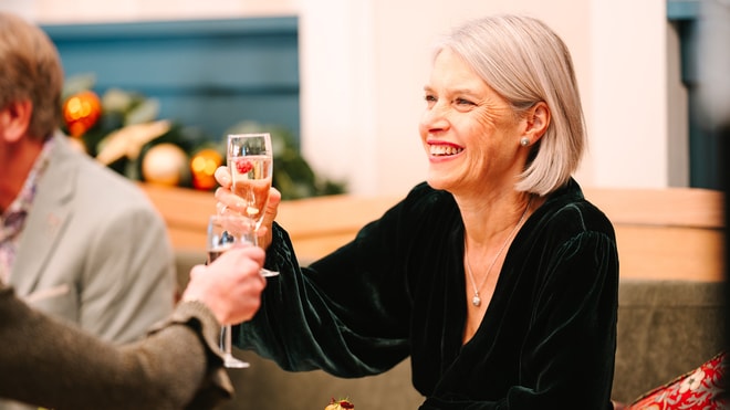 A woman in a black velvet outfit raises a champagne flute while toasting with another person. Festive decorations are visible in the background.