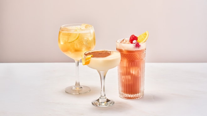 Three colorful cocktails on a marble surface: a bubbly drink with lemon, a creamy one with chocolate, and a fruity mix with raspberries.