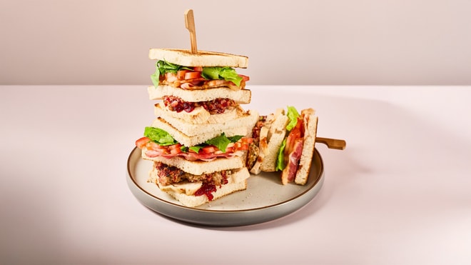 A tall stack of layered sandwiches with lettuce, tomatoes, and meat, served on a round plate against a soft pink background.