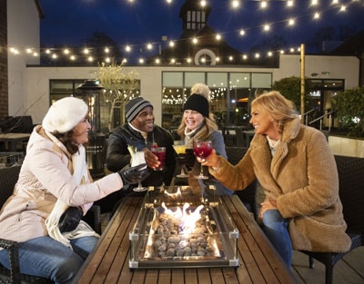 A group of friends enjoy colorful cocktails around a warm fire pit, under twinkling string lights in a cozy outdoor setting.