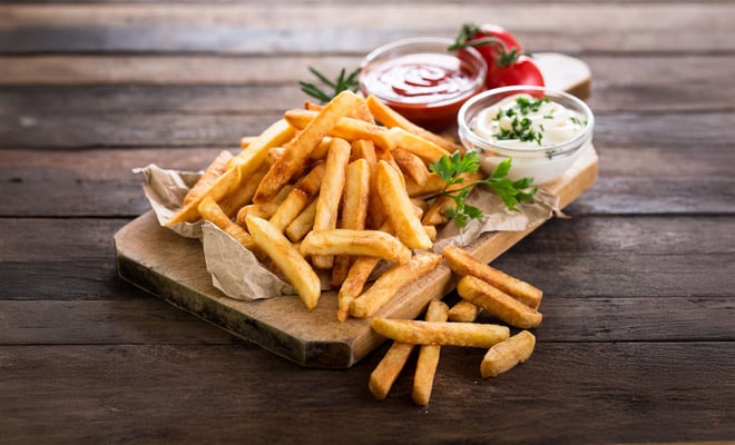 A pile of golden-brown French fries on a wooden board, accompanied by ketchup and mayonnaise in glass bowls, garnished with parsley.