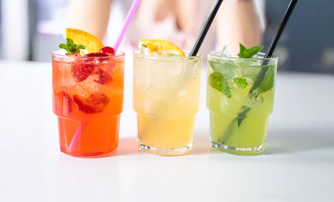 Three colorful cocktails in clear glasses: a red drink with strawberries, a yellow citrus drink, and a green mint-infused beverage.
