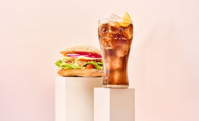 A fresh sandwich with greens, tomatoes, and onions beside a frosty glass of cola with ice and a lemon slice against a soft pink background.