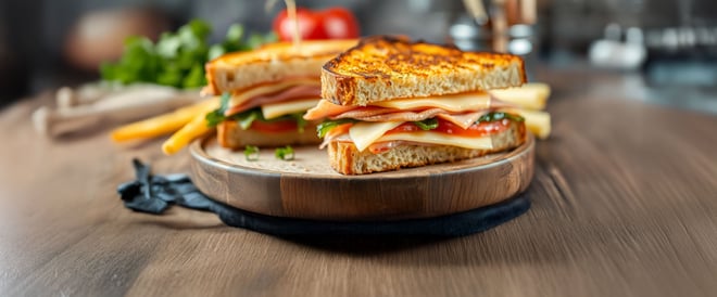 Close-up of a toasted sandwich stacked with ham, cheese, lettuce, and tomato, served on a wooden plate with fries in the background.