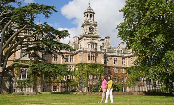 A couple strolls across a lush green lawn in front of a grand historic mansion, adorned with ivy and surrounded by large trees at Thoresby Hall
