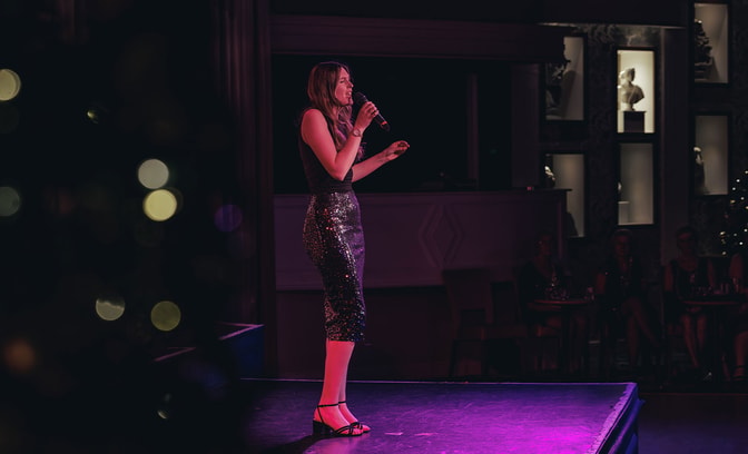 Performer singing on stage under colourful lighting in front of a seated audience.