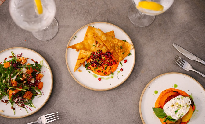 Overhead view of three small plates with colourful dishes, including salad and flatbread with dip, alongside glasses of iced gin and tonics with lemon on a stone table.