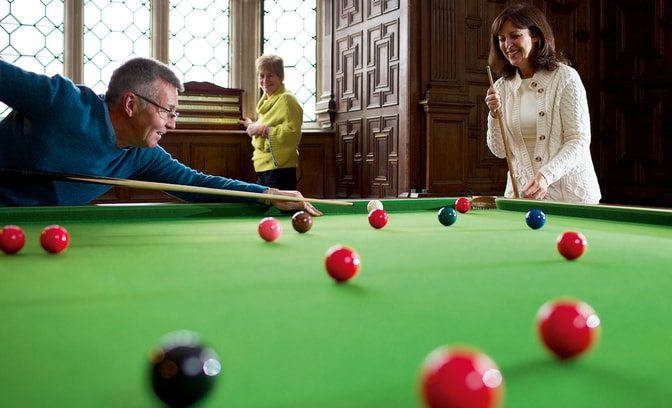 Person taking a shot on a snooker table in a wood-panelled room, with two other people standing nearby. Red and coloured balls are spread across the green baize under natural light from large leaded windows.