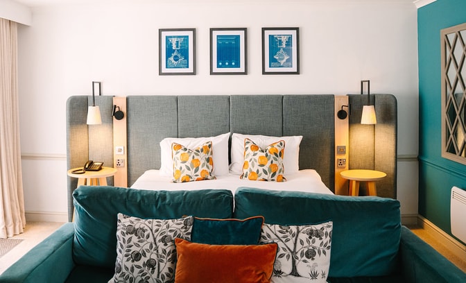 Modern hotel room with a king-size bed, bedside tables and reading lights, complemented by a teal sofa and coordinated accent cushions in a bright, stylish setting.