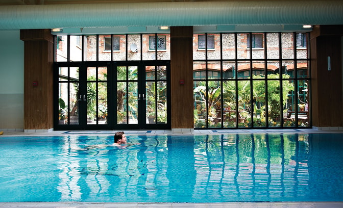 Person swimming in an indoor pool beside floor-to-ceiling windows overlooking a landscaped courtyard. Reflections shimmer across the water, creating a calm and relaxing leisure environment.