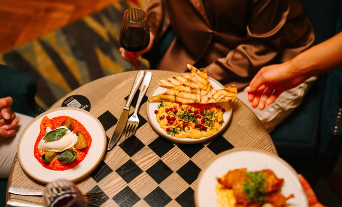 Small plates including flatbread with dip and a burrata-style dish served on a patterned table in the Great Hall, with a glass of red wine and hands reaching to serve food.
