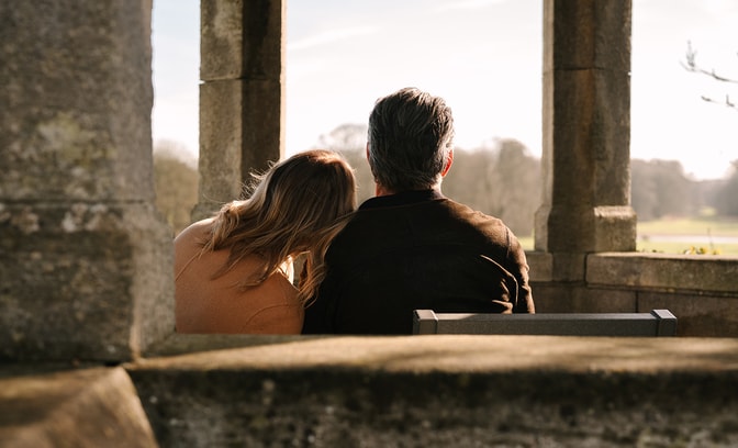 Two people sit closely together on a bench inside a stone structure with tall columns, facing an open landscape of grass and distant trees. One person leans their head against the other’s shoulder. Warm sunlight backlights the scene, creating a soft, quiet atmosphere.