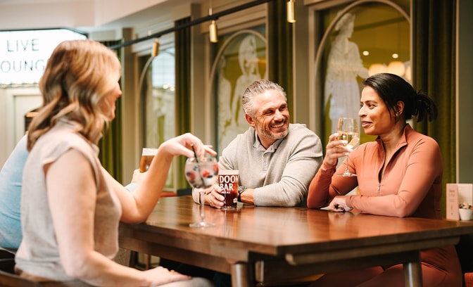Three people sit around a wooden table in a warmly lit lounge, each holding drinks including wine and beer. A sign reading “LIVE LOUNGE” is visible in the background along with framed wall art and hanging lights.
