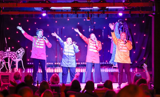 Four members of the Warner Theatre Company at Bembridge Coast performing on stage, with guests waving and joining in
