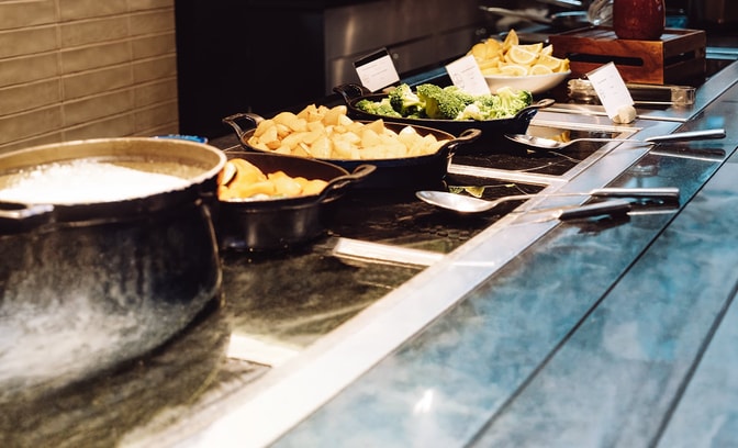 Hot buffet counter in Market Kitchen featuring pots and trays of vegetables, including potatoes, broccoli and lemons, with serving utensils on a modern countertop.
