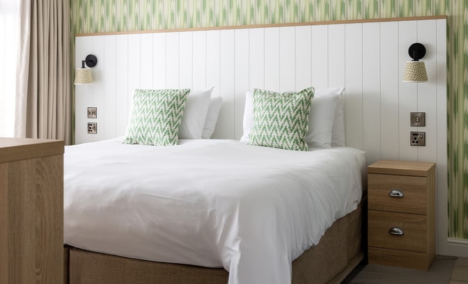A neatly made double bed with white bedding and two green patterned cushions, set against a white paneled headboard and green striped wallpaper, with wall-mounted lamps and wooden bedside furniture.