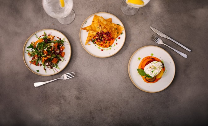 Overhead view of three small plates with colourful dishes, including salad and flatbread with dip, alongside glasses of iced drinks with lemon on a stone table.