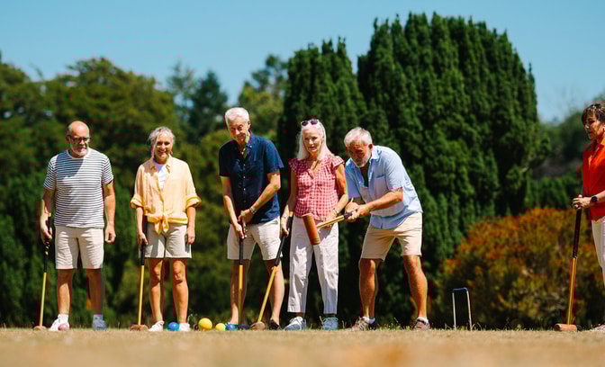 A group of six individuals wearing casual summer clothing stand on a grassy field, preparing to play croquet against a sunny backdrop.