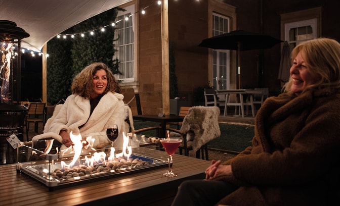 Two women sat in fur jackets by a firepit and enjoying cocktails together