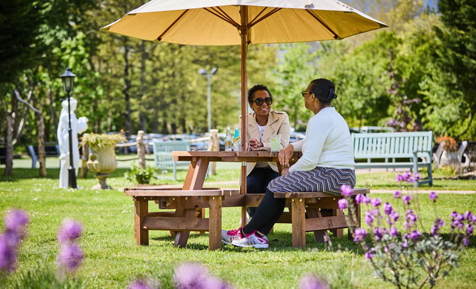 Two women sit at a wooden picnic table under a large umbrella, surrounded by lush greenery and vibrant flowers at Sinah Warren