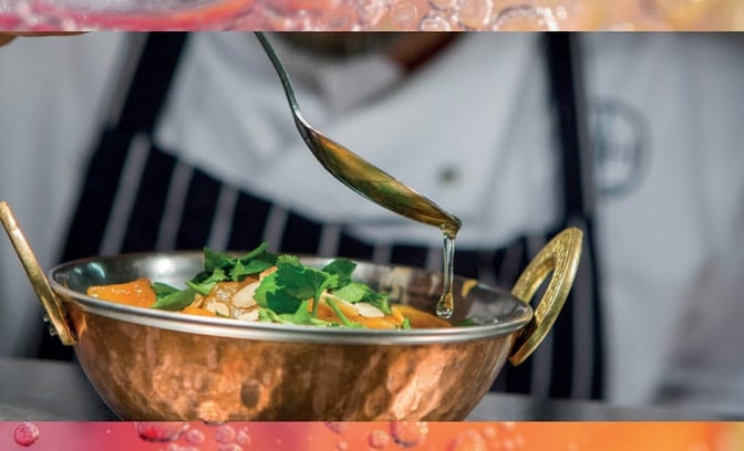 A chef drizzles sauce over a vibrant dish in a decorative copper bowl, garnished with greens and citrus slices.