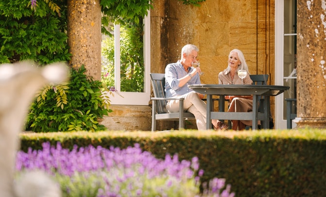 A couple enjoys a relaxed lunch at a garden table surrounded by lush greenery and blooming lavender flowers. Wine glasses are visible.