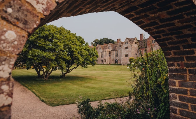 View through an archway of a historic stone at Littlecote house surrounded by a lush green lawn and a vibrant tree in the foreground.