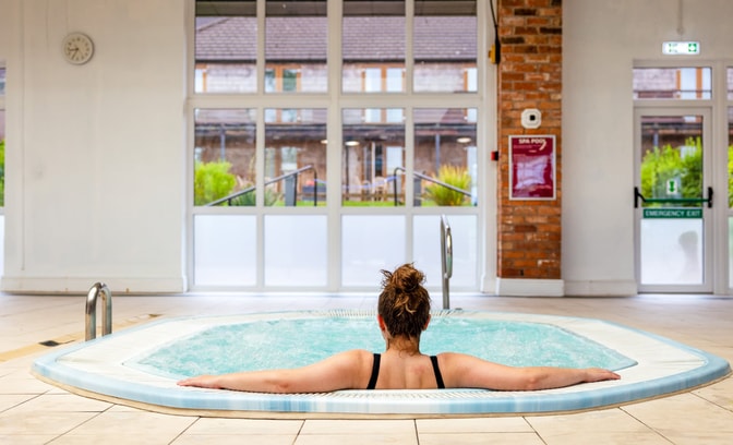 A woman relaxes the bubble pool at Alvaston Hall Hotel, facing large windows that overlook a green garden area. A clock is visible on the wall.