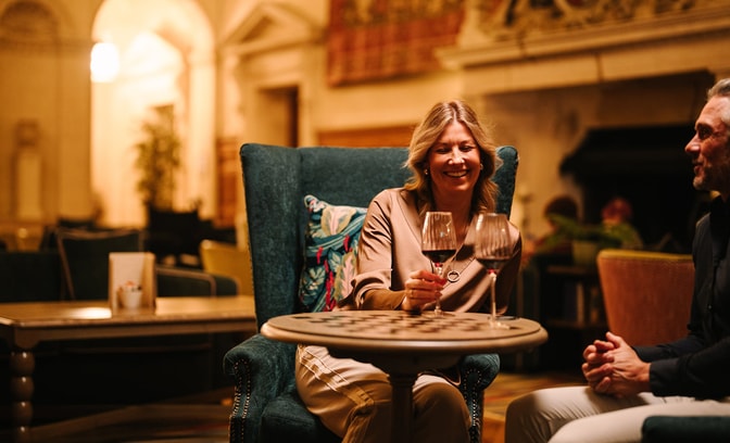 A warmly lit lounge setting with two people seated at a small round table, each holding a glass of red wine. The space features upholstered chairs, patterned cushions, and soft ambient lighting, with blurred seating and architectural details in the background creating a cosy atmosphere.