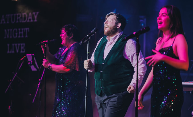 Three singers performing live on stage under colourful lighting, each singing into a microphone.