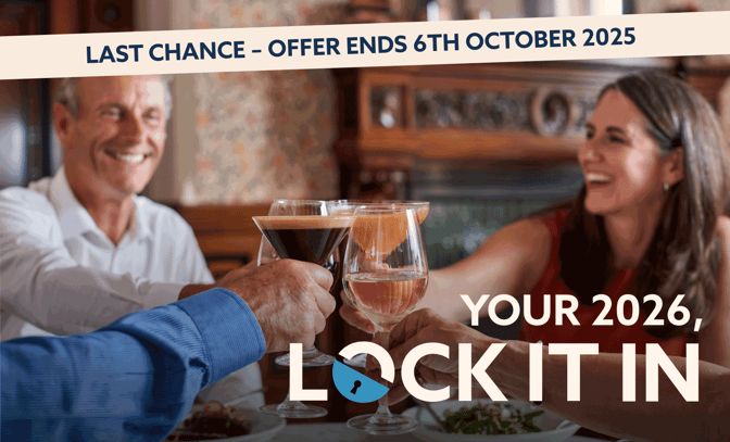 Two pairs of hands clink glasses filled with cocktails at a dining table, with a rustic wooden backdrop and plates of food visible.