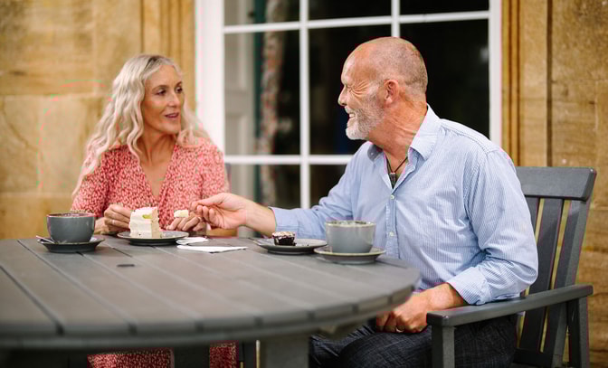 A couple enjoy dessert and coffee at a wooden table outdoors, with a cozy atmosphere and a scenic backdrop.