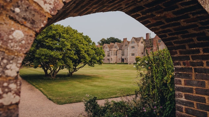 View through an archway of a historic stone at Littlecote house surrounded by a lush green lawn and a vibrant tree in the foreground.