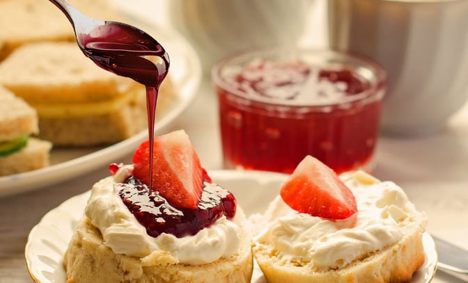 A spoon drizzles red jam over a scone topped with cream and strawberries, with sandwiches in the background.