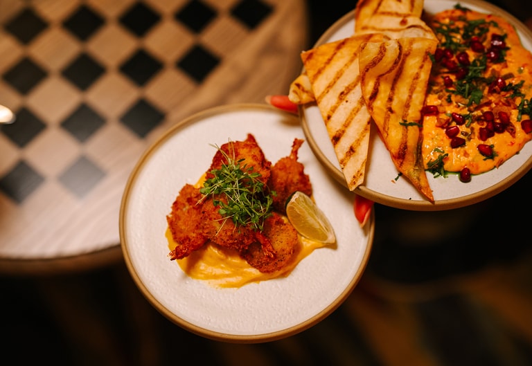 Close-up of small plates in the Great Hall featuring crispy prawns with lemon and flatbread with a spiced dip topped with herbs and pomegranate seeds.