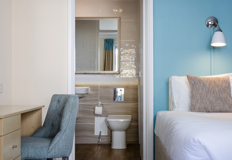 Hotel room with a white bed beside a blue accent wall, next to an open ensuite bathroom featuring a toilet, sink, and mirror, with a desk and upholstered chair in the foreground.