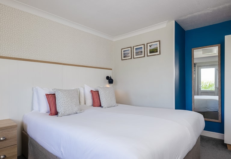 Spacious hotel bedroom with a white bed, patterned and red accent cushions, a textured feature wall, bedside unit, blue accent wall, and framed coastal artwork above.