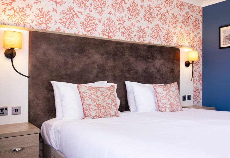 Modern bedroom with a large white bed, coral-patterned accent cushions, a dark headboard, and warm bedside lighting set against a bold patterned feature wall.