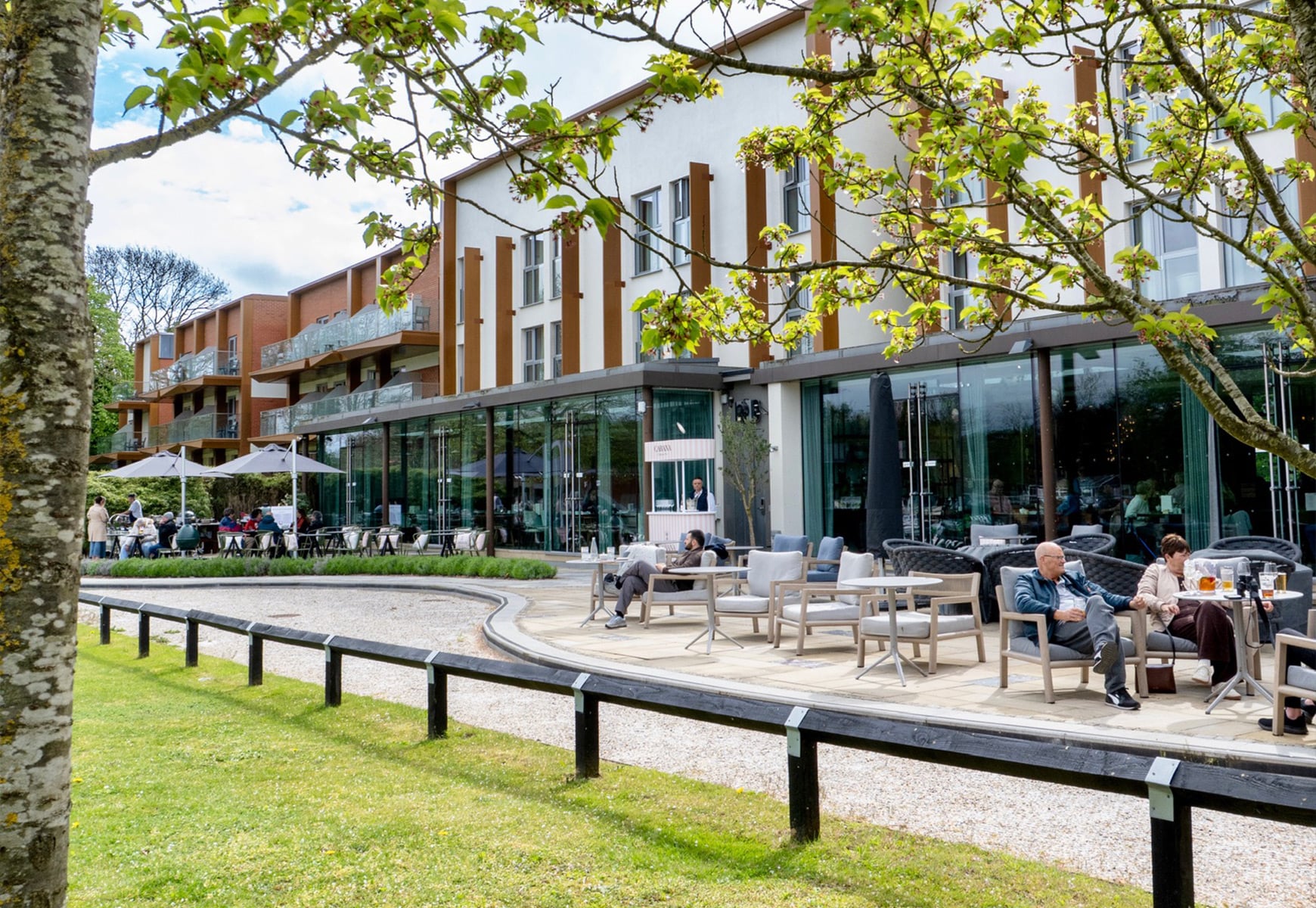 Outdoor terrace with guests seated at tables enjoying drinks and food, set beside a modern building and landscaped gardens.