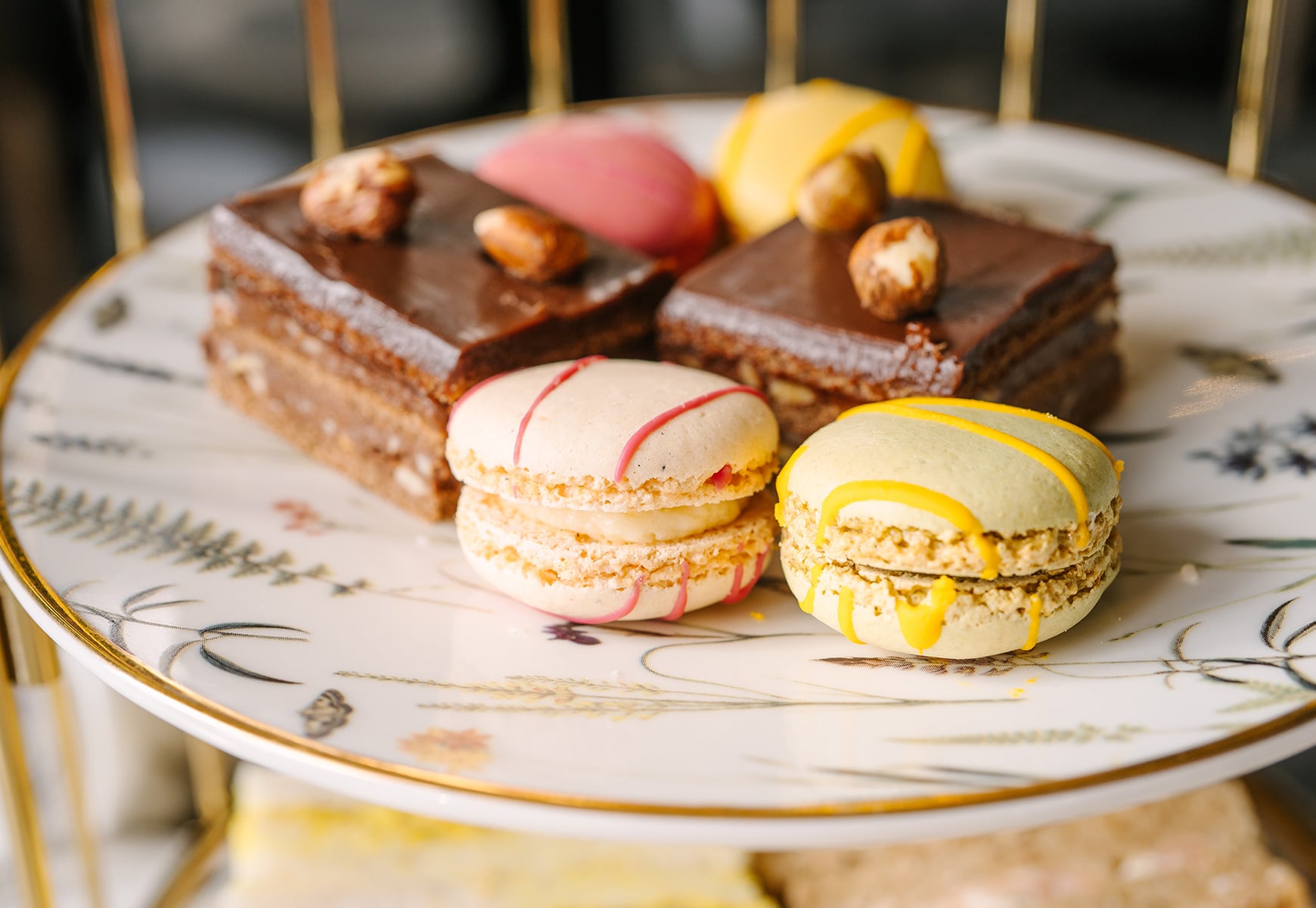 Afternoon tea selection of macarons and chocolate cakes served on a patterned tiered stand in soft, warm lighting.