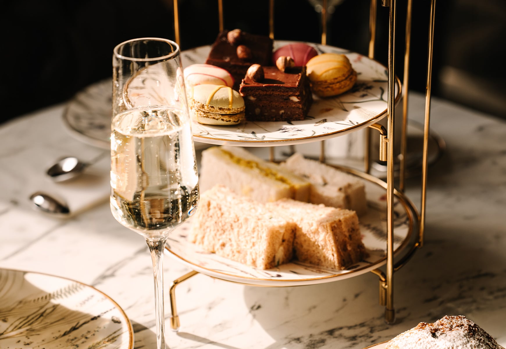 Afternoon tea stand with finger sandwiches, macarons and cakes, served with a glass of sparkling wine on a marble table.