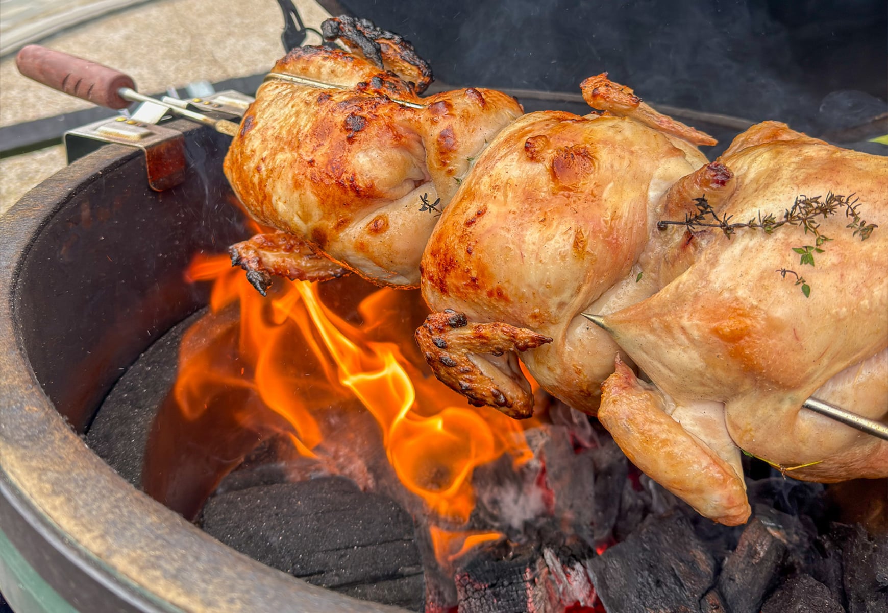 Whole chickens roasting on a Big Green Egg rotisserie above open flames and hot coals.