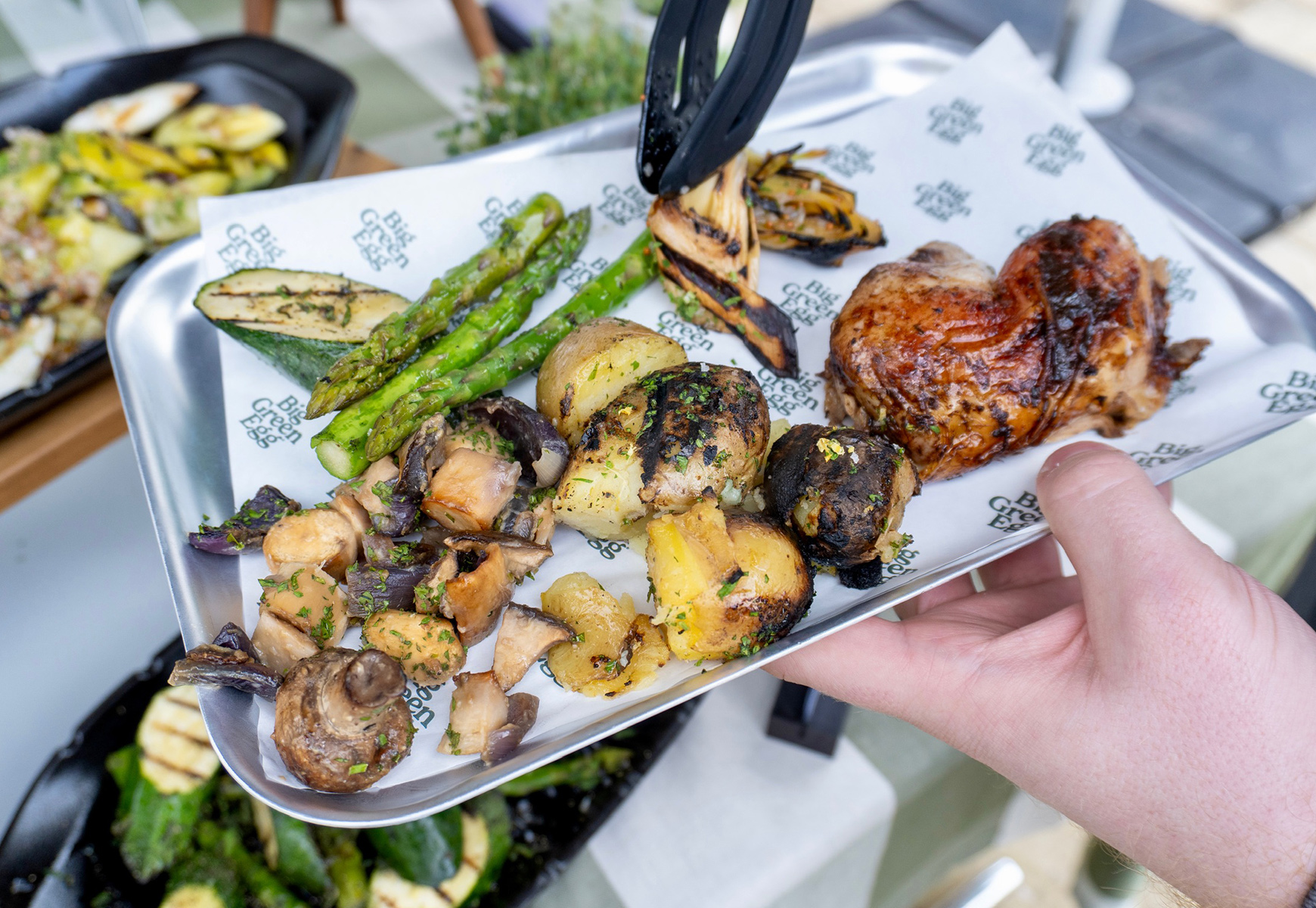 Plate of Big Green Egg food with grilled vegetables, roasted potatoes and chicken being served with tongs at an outdoor station.